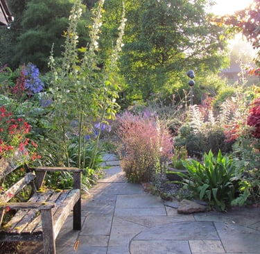 A rustic wooden garden bench sits on a stone patio surrounded by blooming summer flowers and greenery.