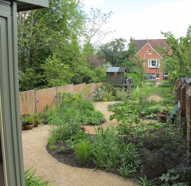 A winding gravel path leads through a lush landscaped backyard garden with flower beds and a wooden playhouse.