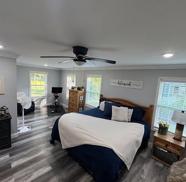 Modern master bedroom with grey LVP flooring, large windows for natural light, and a ceiling fan. 