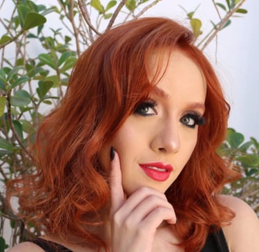 Woman with vibrant copper red hair and red lipstick posing against a green plant backdrop.