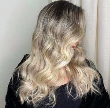 A woman with long wavy blonde balayage hair and dark roots styled in soft beach waves.