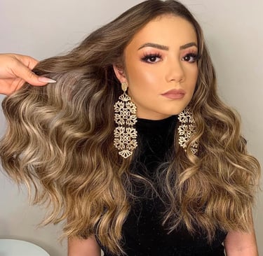 Woman with long wavy balayage hair, glamorous pink eyeshadow, and gold statement chandelier earrings.