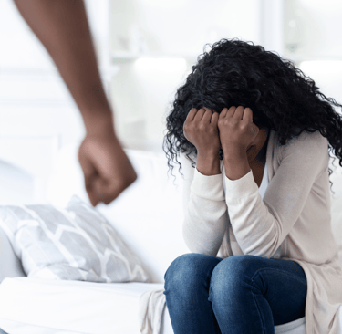 Mujer sentada en su cama abrumada y llorando enfrente de una pareja alterada y agresiva