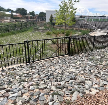 Black decorative metal security fence installed around a landscaped drainage basin with grey river rock.