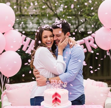 Casal sorridente abraçado em chá revelação de bebê, com balões rosa, confetes e bolo rosa é menina