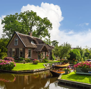 Quaint thatched roof cottage in Giethoorn, Netherlands, with wooden boats on a canal and blooming pink flowers.
