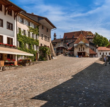 Charming cobblestone street in Gruyères Switzerland with historic buildings and outdoor cafe seating under a blue sky.