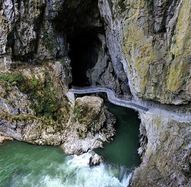 A metal walkway bridge leads into the dark mouth of the Skocjan Caves over a flowing green river.