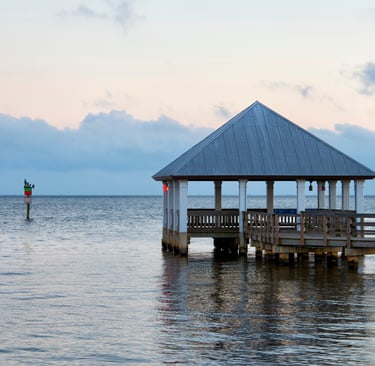 A wooden pier with a gazebo pavilion overlooks a calm ocean bay at sunrise with blue and pink clouds.