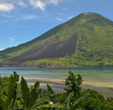A lush tropical volcano with dark lava flows overlooks a turquoise ocean bay under a blue sky.