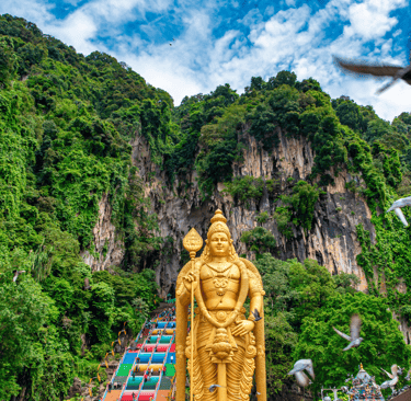 The gold Lord Murugan statue at Batu Caves in Malaysia with limestone hills and pigeons in the foreground.