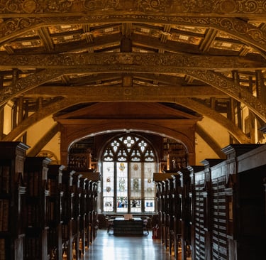 Ornate wooden arched ceiling over rows of bookshelves in a historic library with stained glass windows.