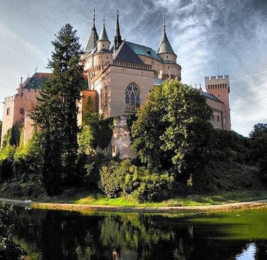 Bojnice Castle in Slovakia reflecting in a lake surrounded by lush green trees under a cloudy sky.