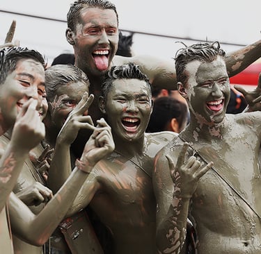 A group of friends covered in mud laughing and gesturing at a mud festival or obstacle course race.
