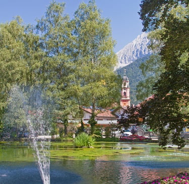A scenic park in Mittenwald, Germany featuring a pond with a water fountain and the Alps in the distance.