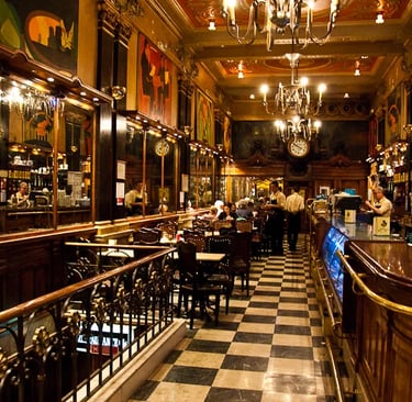 Historic European cafe interior featuring checkered floors, ornate gold accents, and a classic wooden bar.