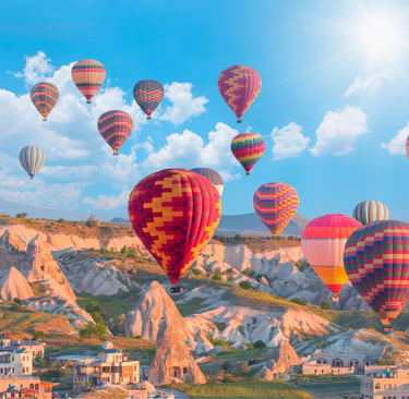 Colorful hot air balloons flying over fairy chimneys and rocky valleys in Cappadocia, Turkey, under a sunny sky.