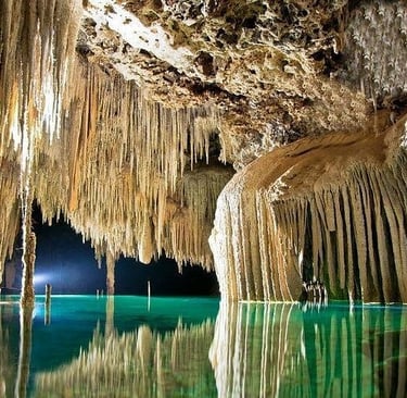 Underground limestone cavern featuring sharp stalactites reflecting in a clear turquoise cenote pool.