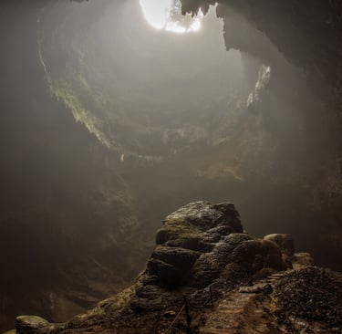 Sunlight streams through a hole in the ceiling of a dark limestone cave onto a central rock formation.