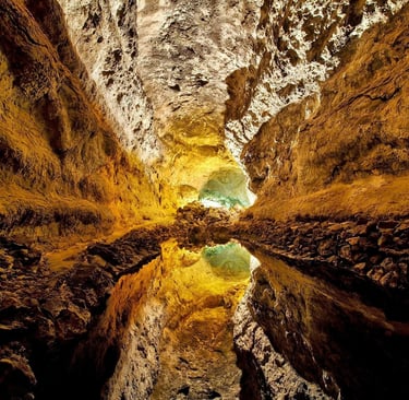 Inside an ancient volcanic cave with a reflective pool on the floor and warm light illuminating the rock formations.