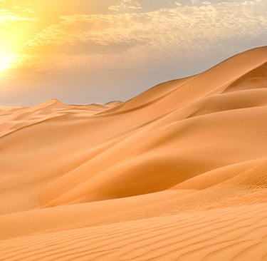 Golden sunset over rolling sand dunes in a vast desert landscape with a glowing sun and hazy sky.