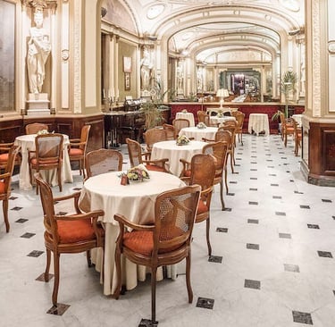 Elegant hotel dining room with marble floors, white statues, and round tables with orange chairs.