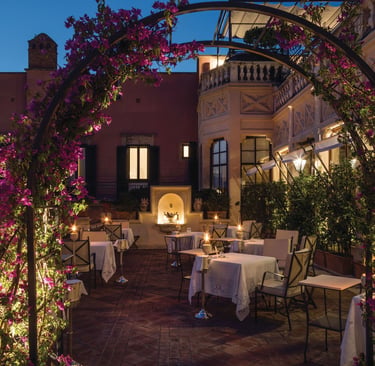 Luxury terrace restaurant in Italy at night with white tablecloths and a purple bougainvillea flower arch.