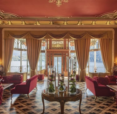 Elegant hotel lounge with red velvet sofas and a panoramic view of Lake Como through large windows.