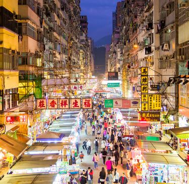 Crowded night market in Hong Kong with glowing neon signs and street food stalls between high-rise buildings.