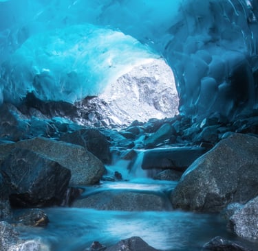 A flowing stream through a glowing blue glacial ice cave with large rocks and a mountain view.