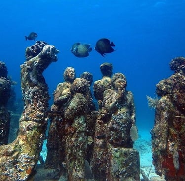 Underwater statues covered in coral reef at an ocean sculpture park with tropical fish swimming nearby.