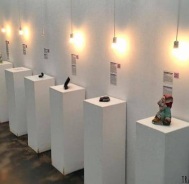 Modern art gallery exhibition featuring small sculptures on white pedestals under hanging pendant lights.