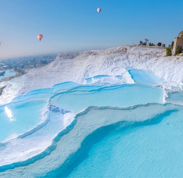 Colorful hot air balloons flying over the blue thermal pools and white travertine terraces of Pamukkale, Turkey.