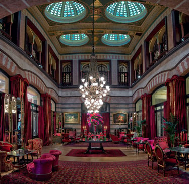 Elegant hotel lounge with ornate stained glass domes, a crystal chandelier, and red velvet armchairs.