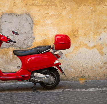 A classic red Italian motor scooter parked against a rustic, yellow textured wall on a cobblestone street.