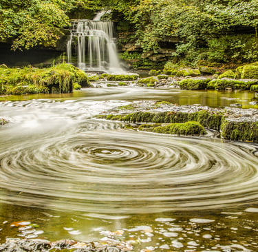 Long exposure of a swirling water vortex in a river below a scenic forest waterfall.