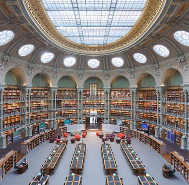 The historic circular reading room at the Richelieu Library in Paris featuring a glass dome and tiers of bookshelves.