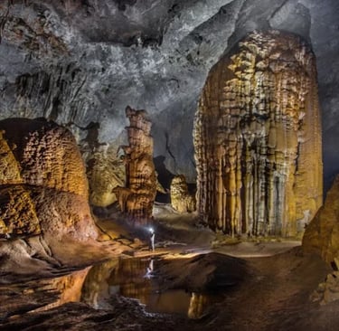 A cavern explorer with a headlamp stands amidst massive limestone stalagmites and rock formations inside a dark cave.
