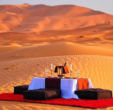 A romantic luxury dinner setup for two on a red rug among the Sahara Desert sand dunes at sunset.