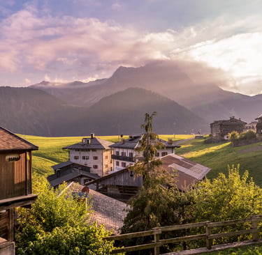 Sunset over a rustic alpine village in the Dolomite Mountains with wooden chalets and green hills.