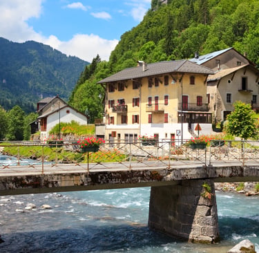 A stone bridge with flowers spans a turquoise river in a scenic French mountain village.