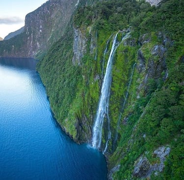 Aerial view of a tall waterfall cascading down lush green cliffs into the blue waters of Milford Sound, New Zealand.