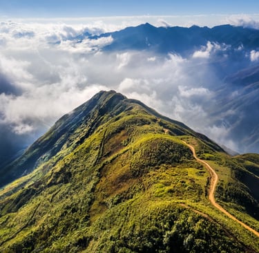 Aerial view of a winding dirt path on a green mountain ridge surrounded by clouds and sunlit peaks.
