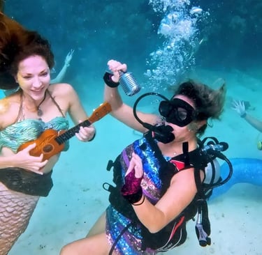 Mermaid and scuba divers performing a concert underwater with a ukulele and microphone on the ocean floor.
