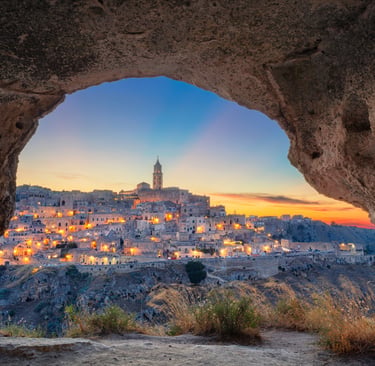 Illuminated ancient city of Matera, Italy, at sunset viewed through a stone cave opening.