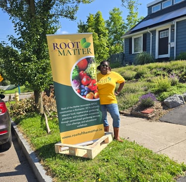 A woman in a yellow Root of the Matter t-shirt stands by a tall banner for community gardening in Rochester, NY.