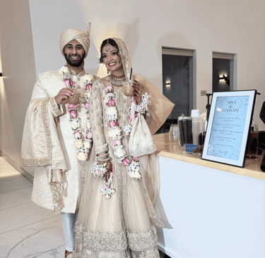 A smiling bride and groom in traditional ivory wedding attire posing with drinks at their reception.
