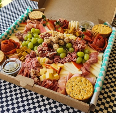 Gourmet charcuterie box with assortment of cured meats, cheeses, grapes, and crackers on a checkered tablecloth.