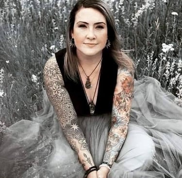 A woman with intricate sleeve tattoos sits in a flower field wearing a tulle dress and bohemian jewelry.
