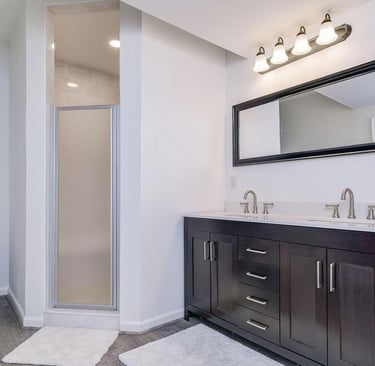 En-suite with custom shower, toilet, and a dual sink vanity. Grey LVT flooring, dark wood vanity.